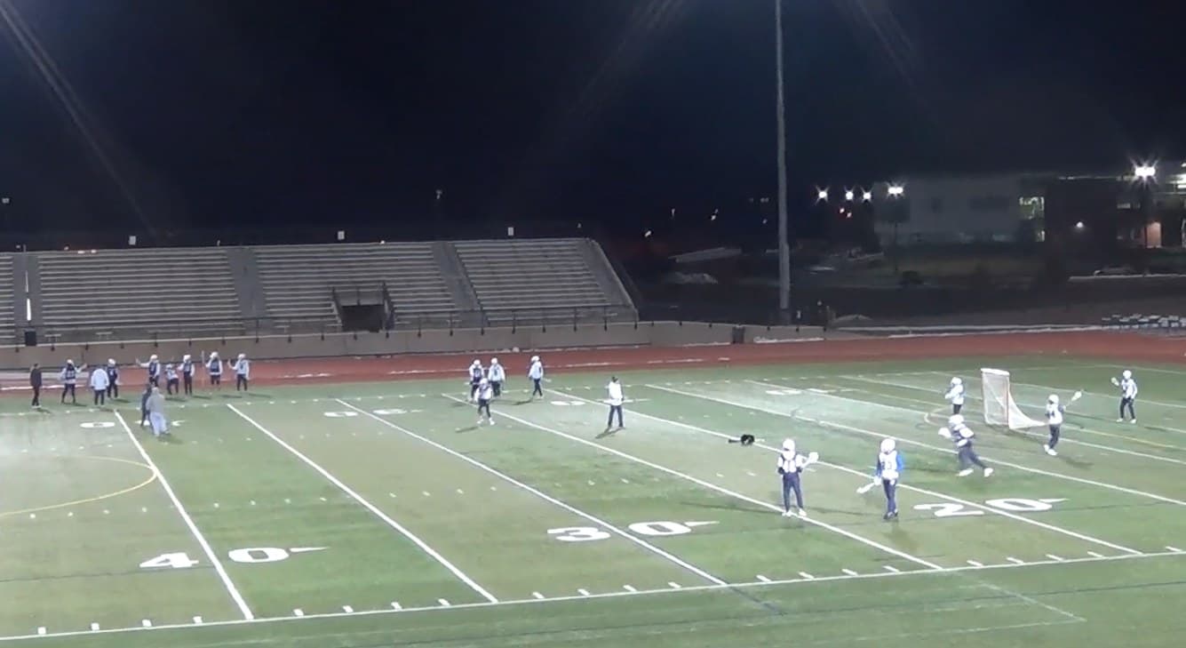 A night lacrosse practice under stadium lights: coaches on the field running separate groups of players across the 40, 30 and 20 yard lines, players moving in each group